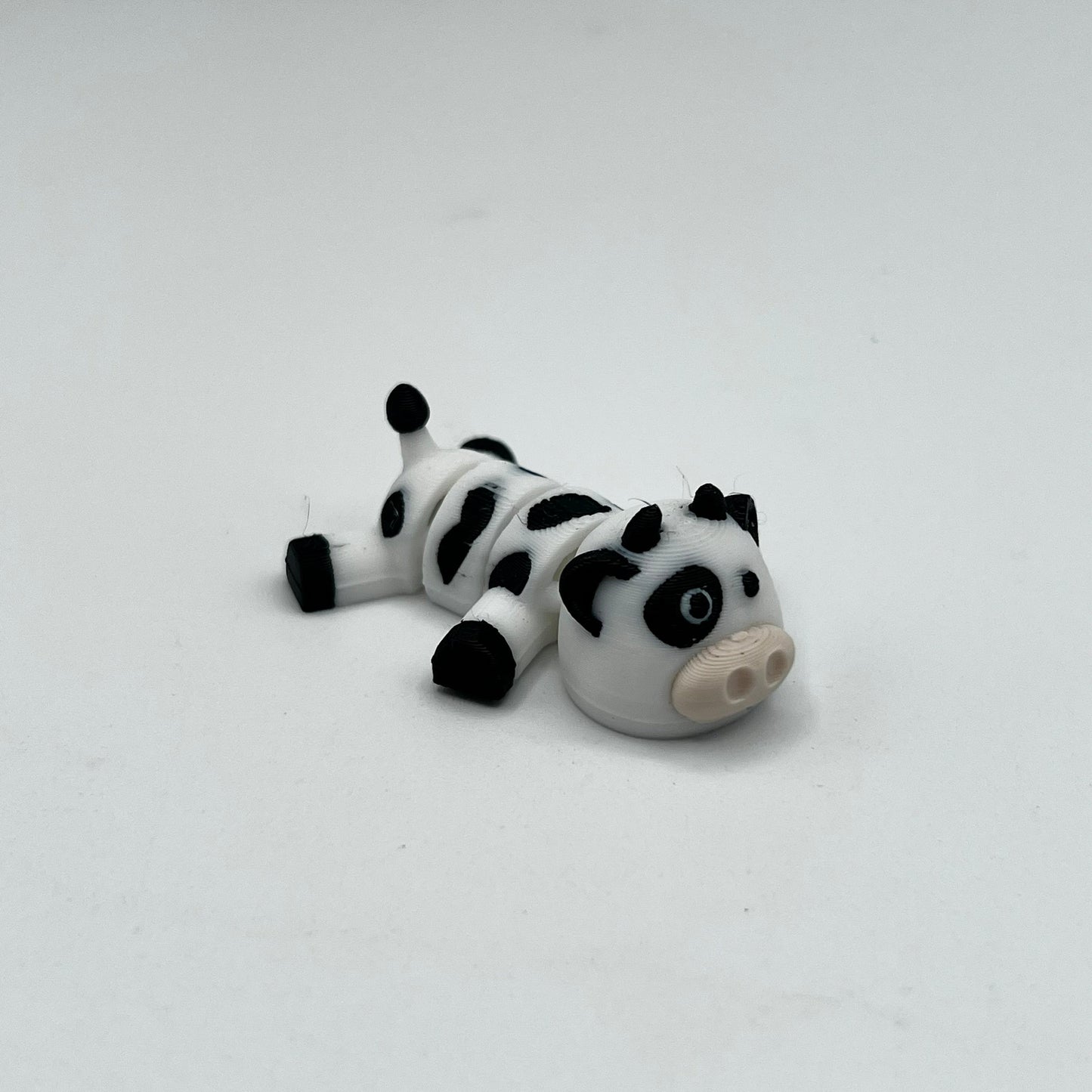 A small black and white spotted cow figurine lying on its side, photographed on a plain light gray background.