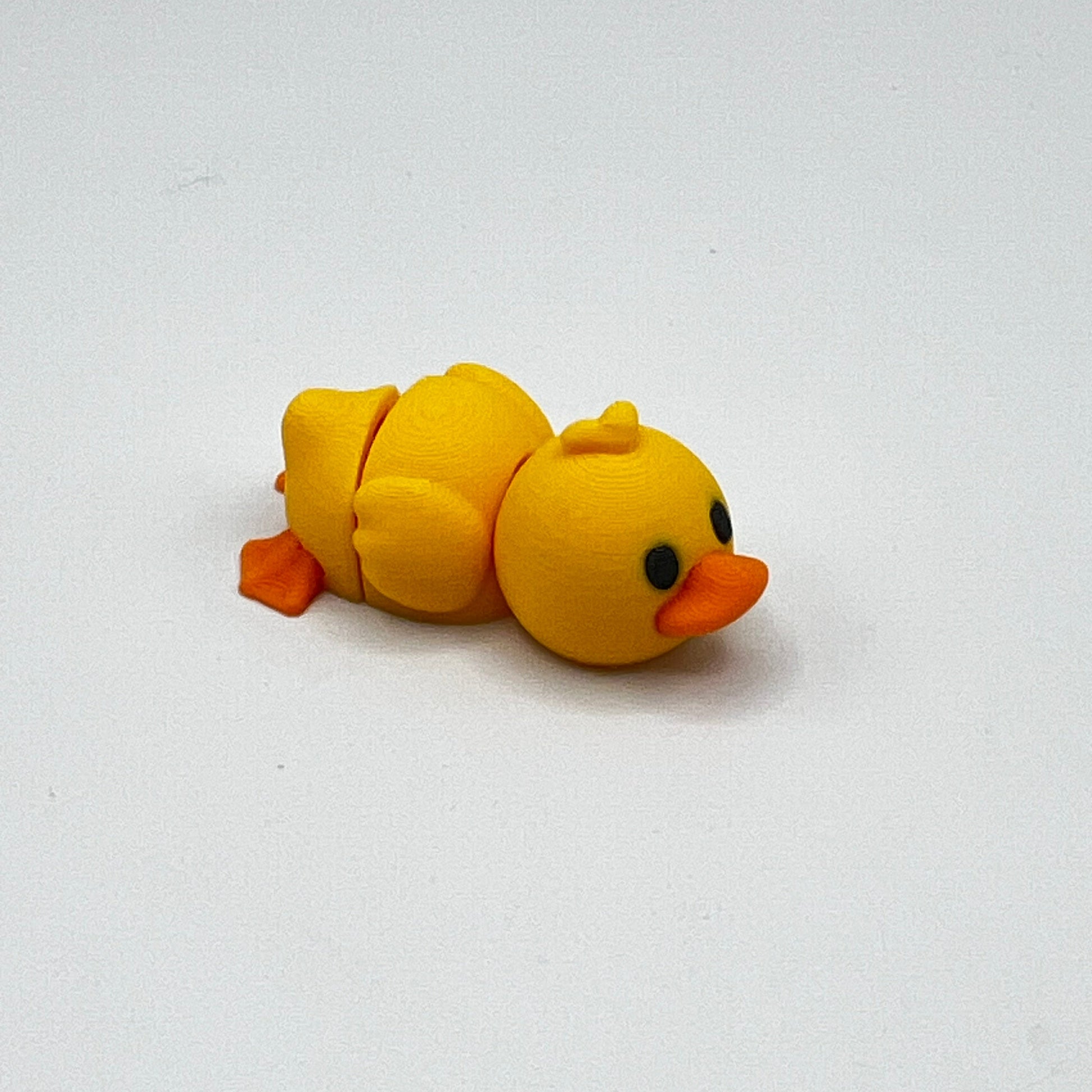 A small yellow duck figurine lying on its side with an orange beak and feet, photographed on a plain light background.