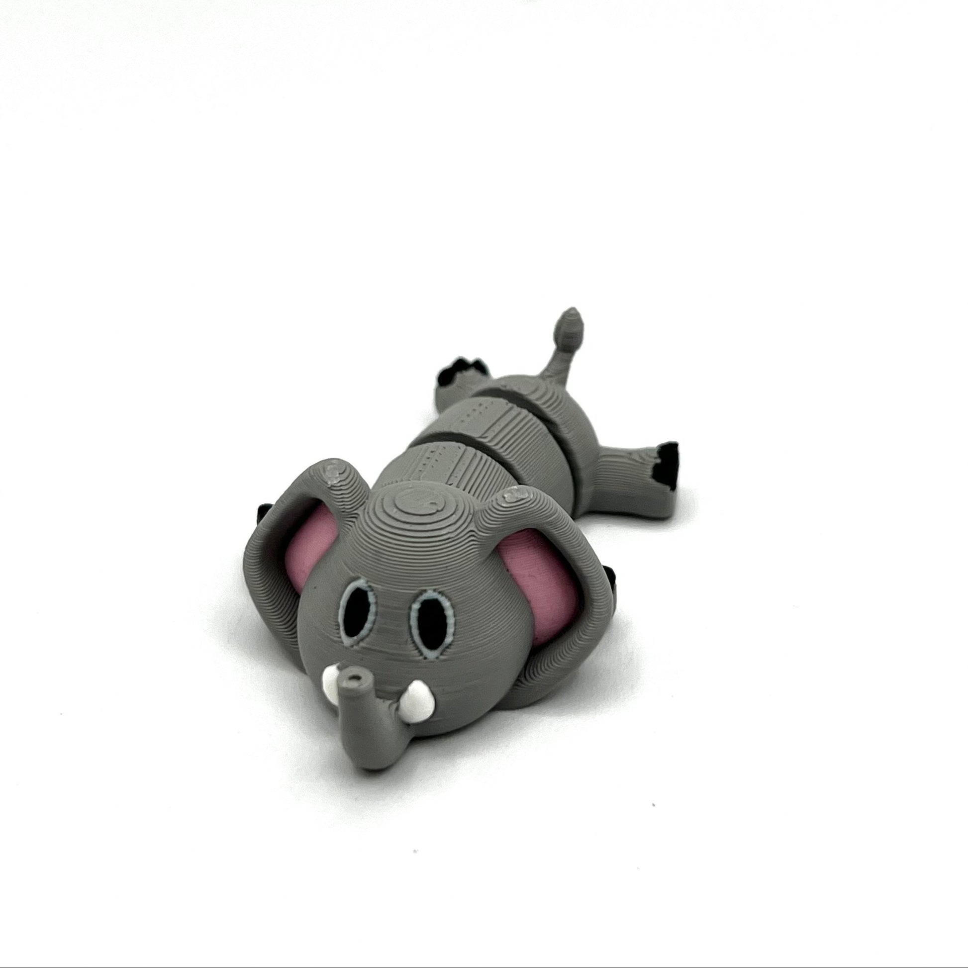 A small gray elephant figurine lying on its side, with pink inner ears, black feet, and white tusks, photographed on a plain light gray background.