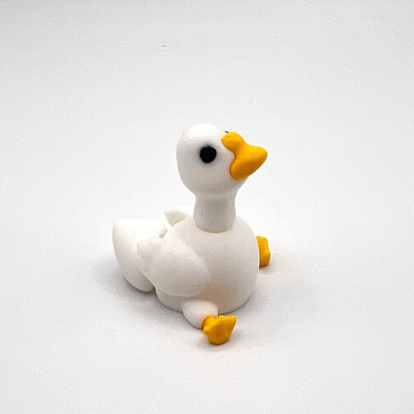 A small white duck figurine with a yellow beak, yellow feet, and a black eye, sitting on a plain light gray background.