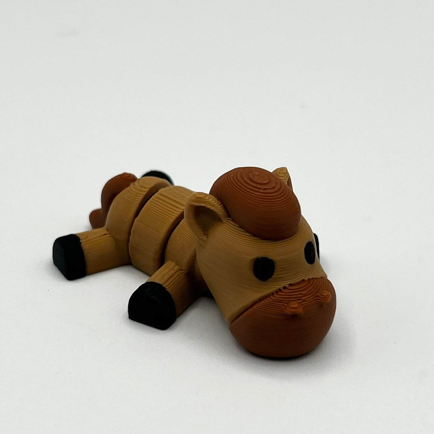 A small 3D-printed toy horse figurine in shades of brown and tan with black hooves, lying down on a plain white background.