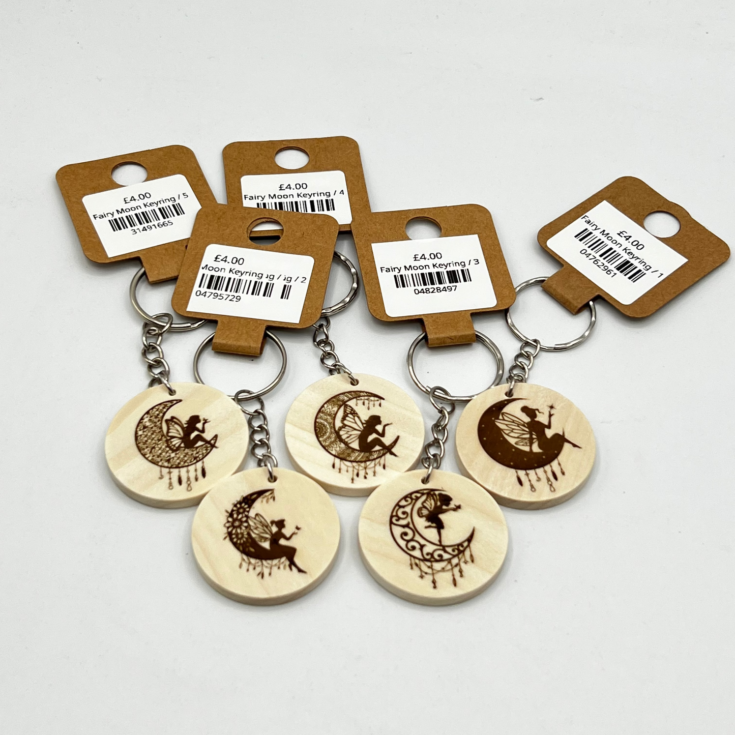 Five small wooden keychains with engraved fairy and moon designs, each attached to a brown paper hang tag with a barcode label, photographed on a light gray background.