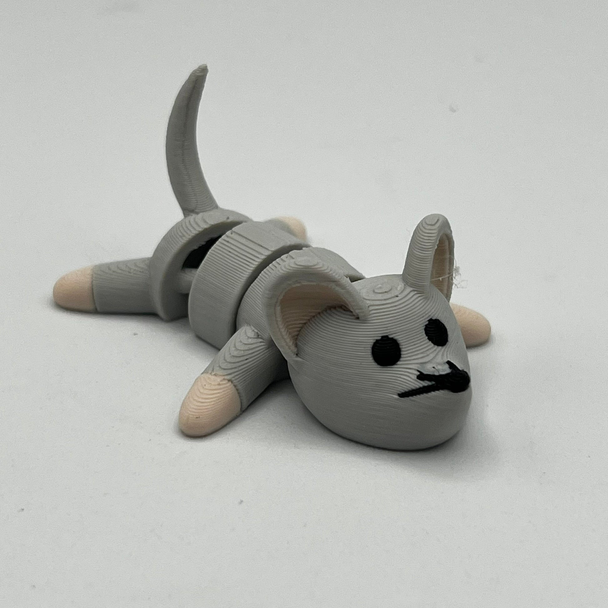 A small gray 3D-printed articulated mouse figurine lying on its side, featuring black printed eyes, nose, and mouth details, with beige paws and tail tips, set against a plain light gray background.