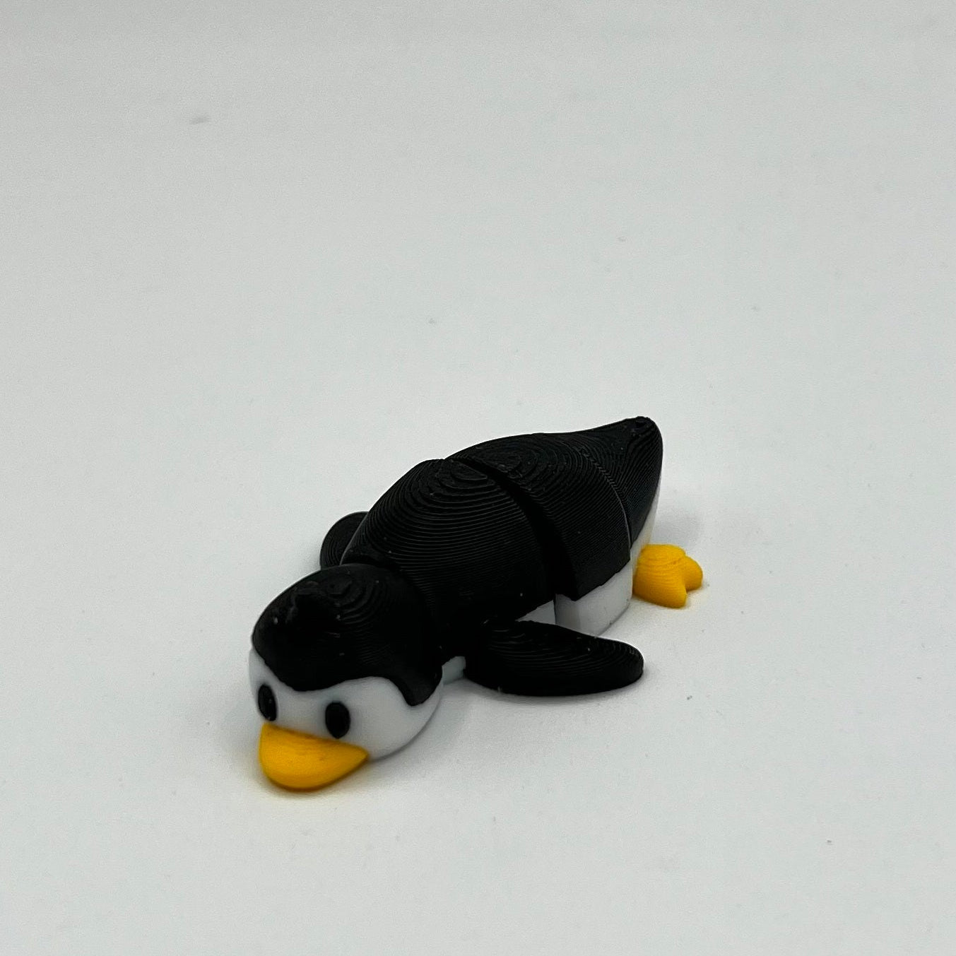 A small penguin figurine with a black and white body and yellow beak and feet, lying on a plain light gray surface.