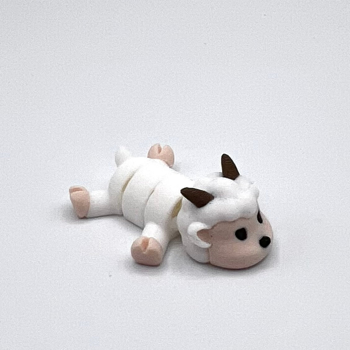 A small white sheep figurine lying on its side, with dark brown horns, black eyes, and pinkish-tan hooves and face, photographed on a plain light gray background.